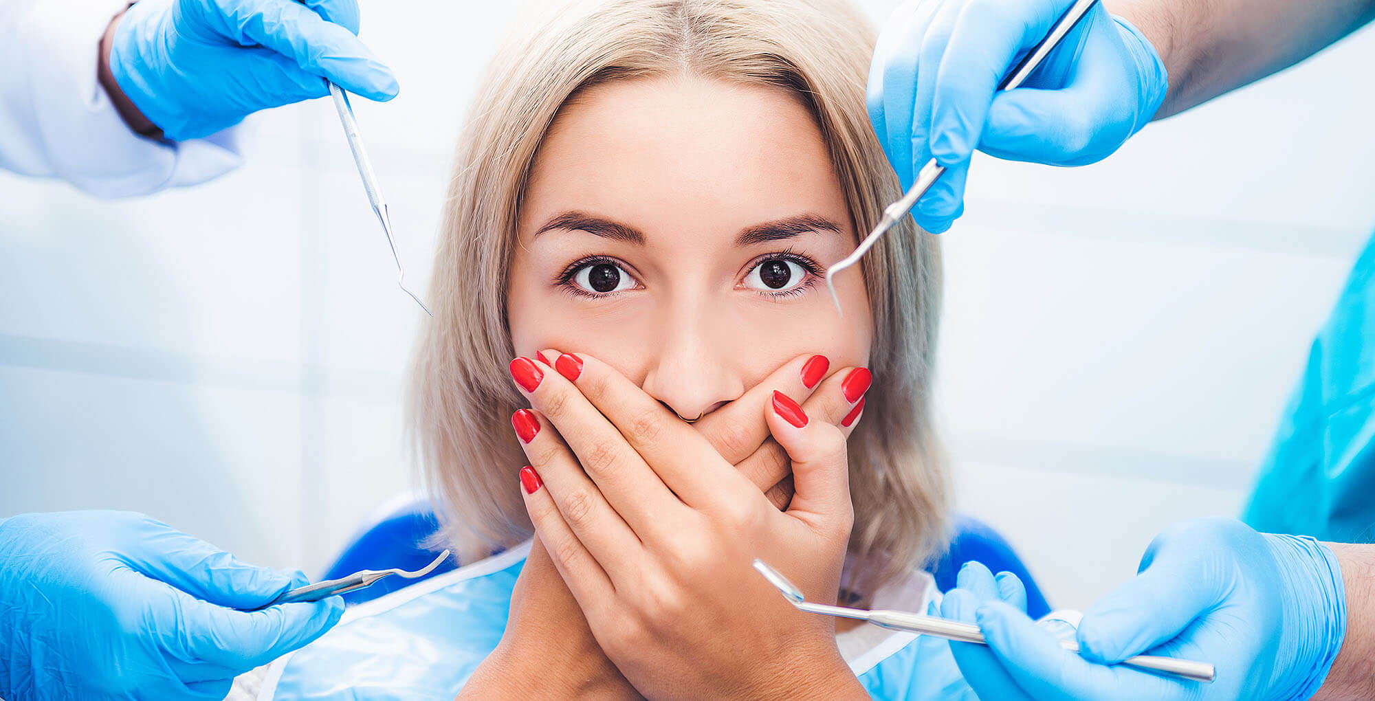 Eine blonde Frau mit ängstlichem Gesichtsausdruck hält ihre Hände vor den Mund. Um sie herum sind mehrere zahnärztliche Instrumente zu sehen, die von Händen in blauen Handschuhen gehalten werden. Die Szene visualisiert Zahnarztangst.