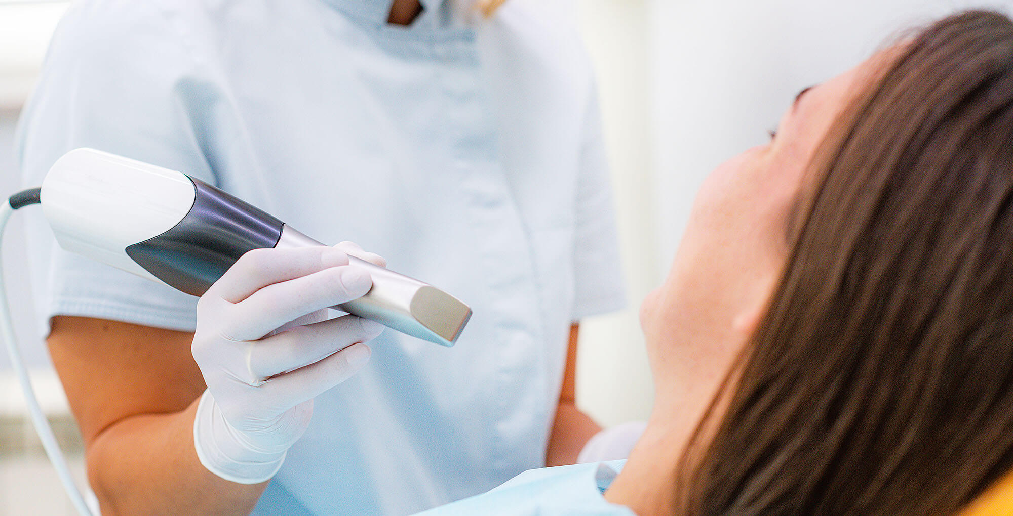 Eine Zahnärztin in weißer Kleidung und weißen Handschuhen verwendet einen Intraoralscanner, um einen digitalen Abdruck vom Gebiss einer Patientin zu nehmen. Die Patientin liegt entspannt im Behandlungsstuhl. Die Szene zeigt eine moderne Methode der Zahnerfassung.