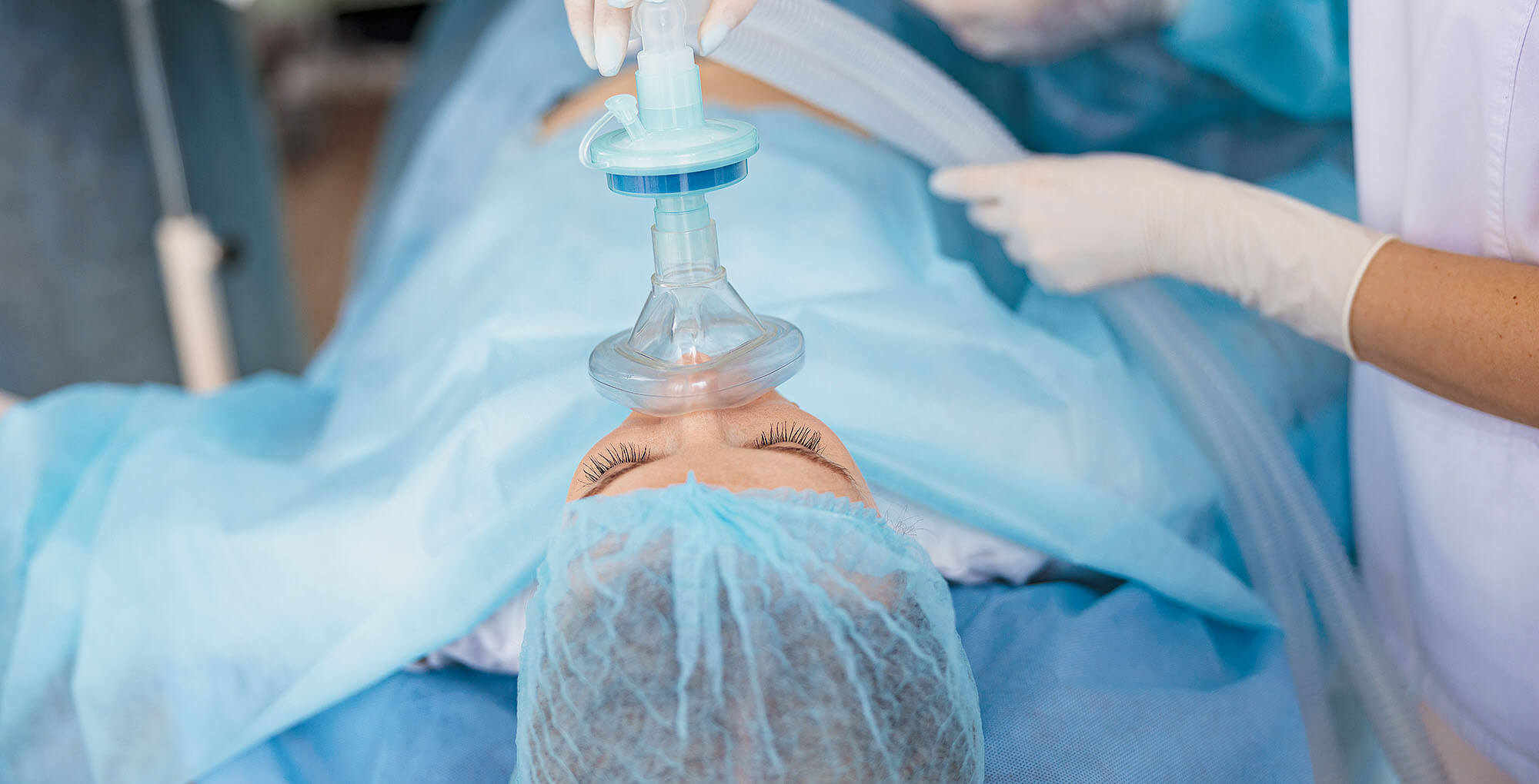 Eine Patientin liegt auf einem Operationstisch, trägt einen blauen OP-Kittel und eine Haube. Sie bekommt über eine Beatmungsmaske Narkose verabreicht. Eine medizinische Fachkraft mit Handschuhen hält die Maske.
