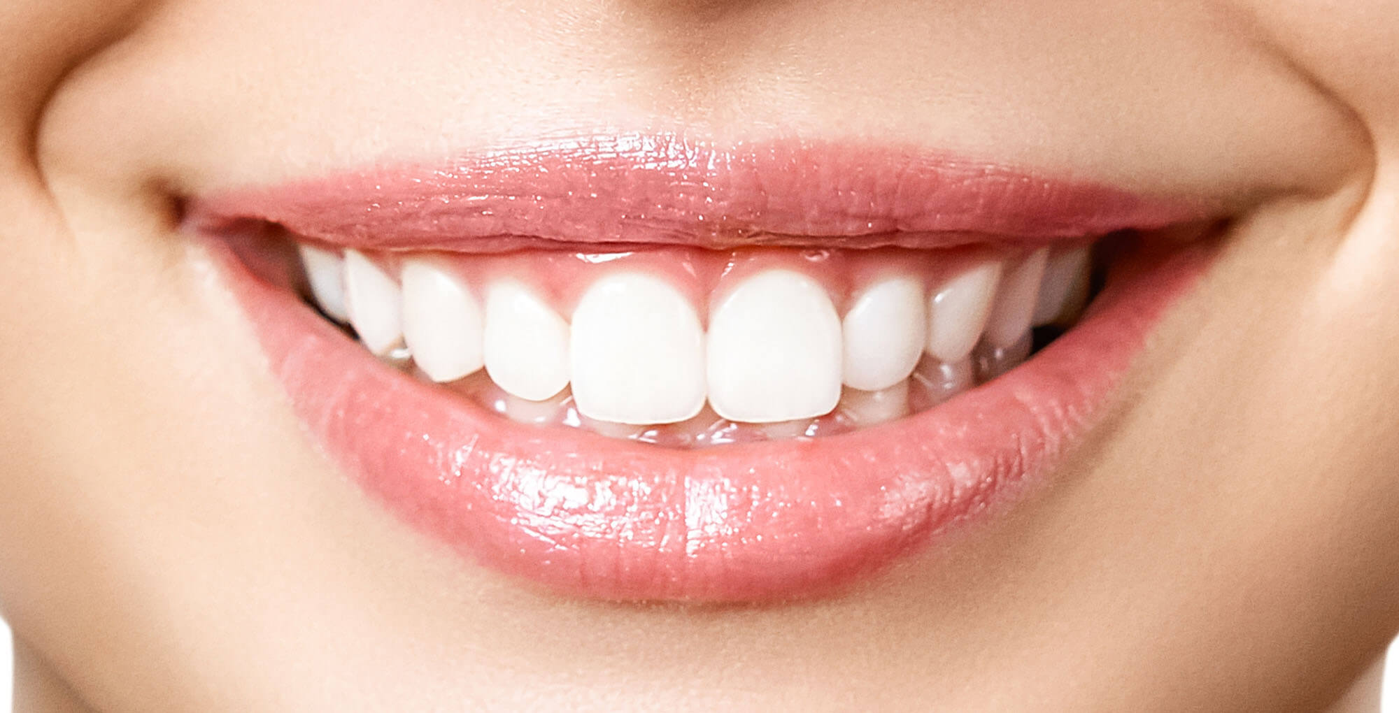 Eine extreme Nahaufnahme zeigt einen lächelnden Mund. Die Lippen sind voll und glänzend mit einem hellrosa Farbton. Die oberen und unteren weißen Zähne sind gut sichtbar und zeigen ein freundliches Lächeln. Die umgebende Haut ist weich und leicht getönt.