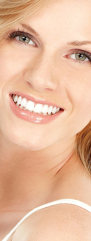 Eine Nahaufnahme zeigt das Gesicht einer lächelnden jungen Frau mit blonden Haaren und hellen Augen. Ihr Mund ist leicht geöffnet und zeigt eine Reihe weißer, gerader Zähne. Ihre Lippen sind voll und glänzend. Ein Teil ihres weißen Oberteils ist im unteren Bildbereich sichtbar.