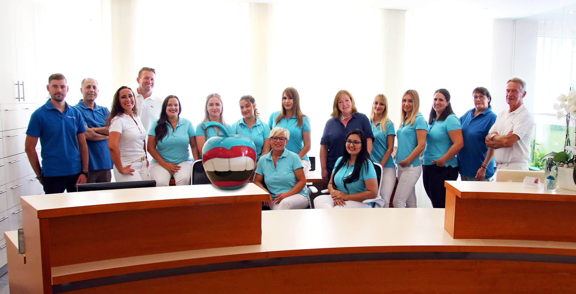 Ein Gruppenfoto zeigt das freundliche Team einer Zahnarztpraxis in Saarbrücken. Mehrere Frauen und Männer in blauen und weißen Oberteilen stehen oder sitzen hinter einem hellen Empfangstresen. Der Hintergrund ist hell und modern.